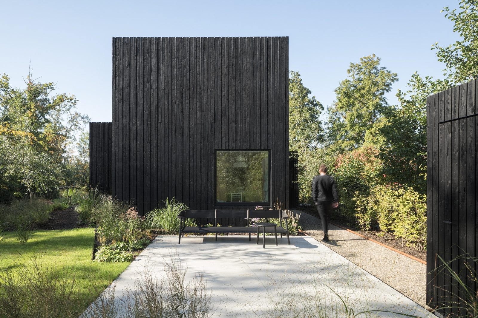Zwart houten kubusvormig huis met brede beton inrit en natuurlijke plantenborder