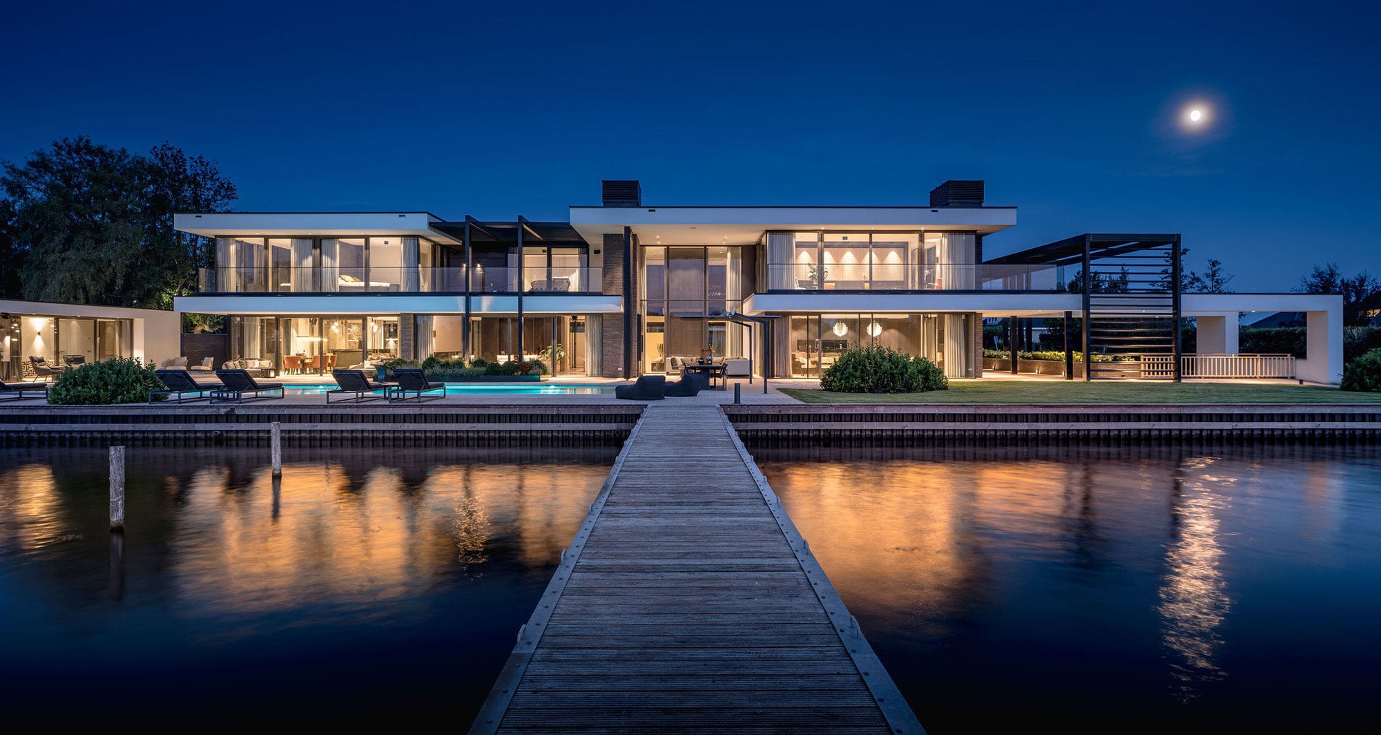 Waterfront villa met houten steiger en gevelverlichting tegen blauwe avondhemel