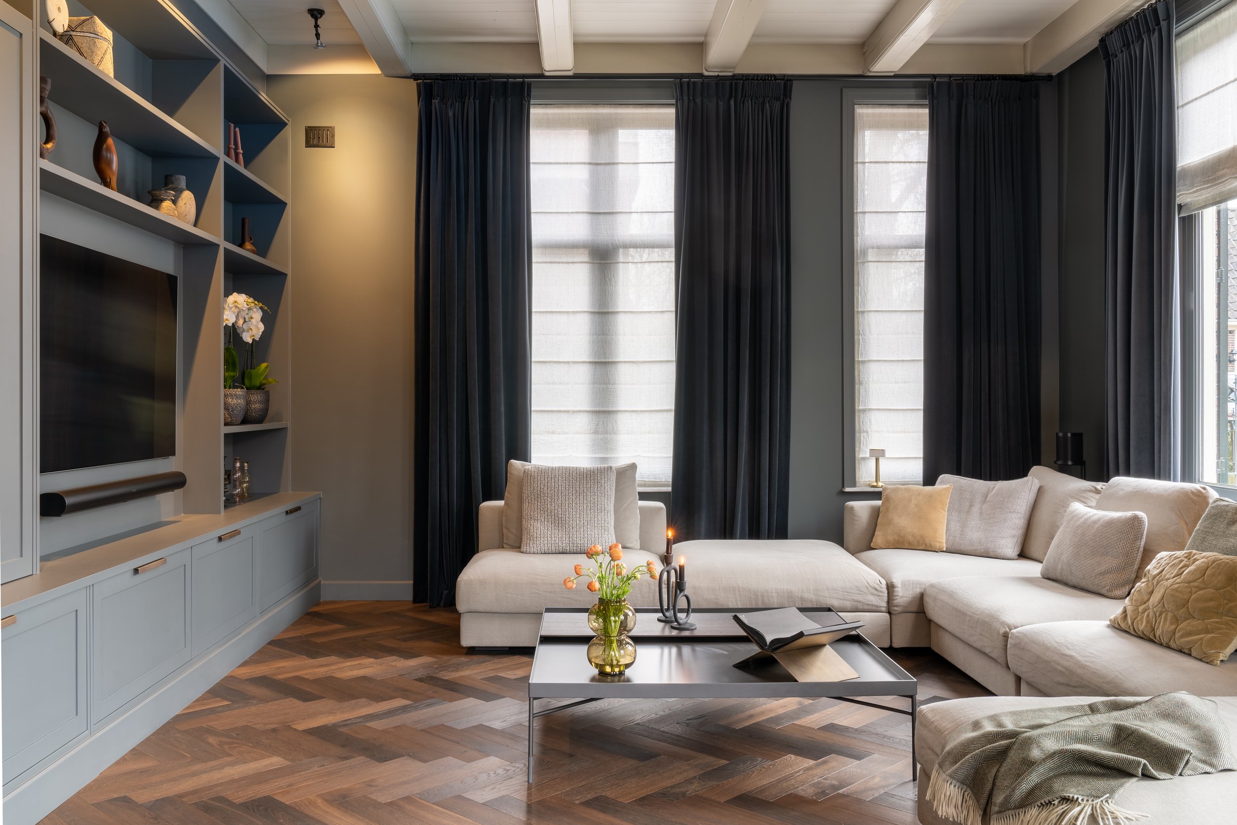 Woonkamer met grijze inbouwkasten, beige hoekbank en zwarte gordijnen